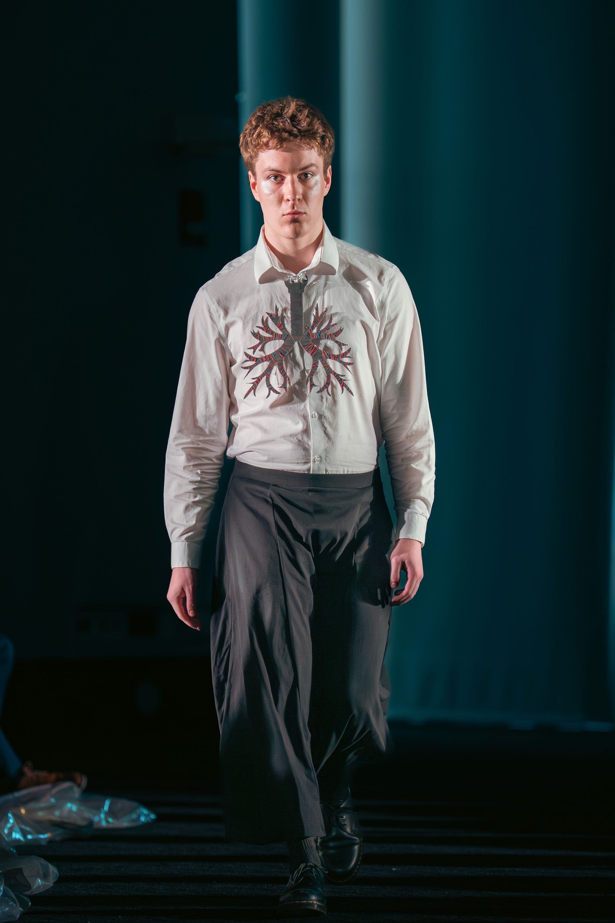 Model walks down the runway in a white button-down shirt embroidered with a stylized bronchial tree motif across the chest in red and black thread. He pairs the shirt with wide-legged black trousers and black shoes.